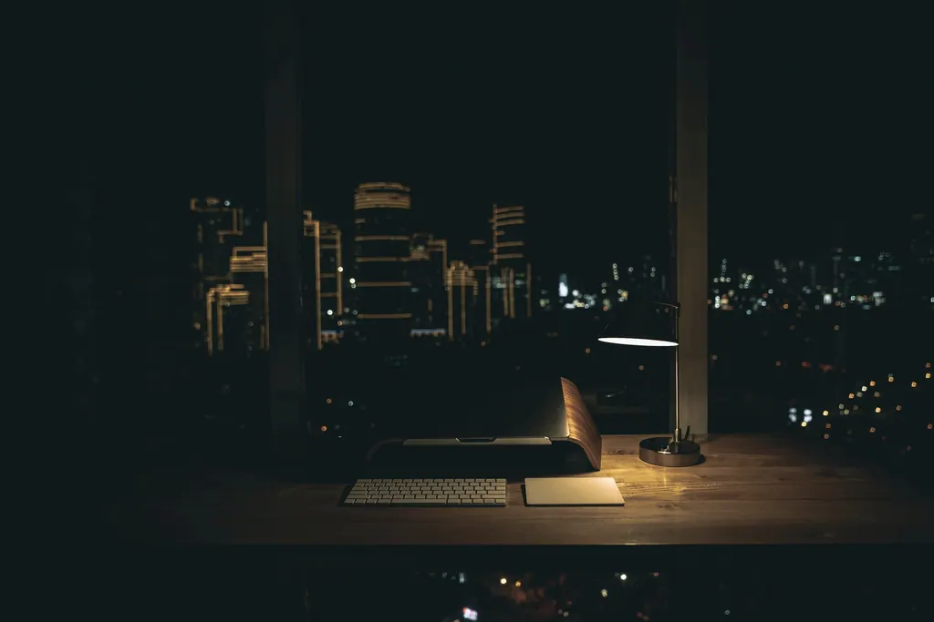 Mesa de trabalho iluminada por um abajur diante de uma janela com a cidade à noite.